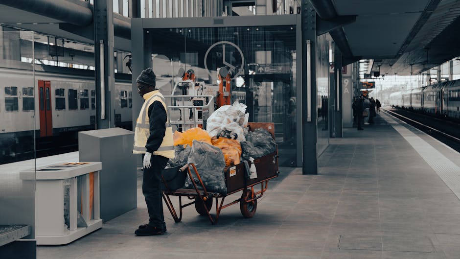 A man dressed in dark clothing, a grey beanie, and white gloves stands on a modern train station platform next to a large red hand truck loaded with various bags of rubbish, including black and orange trash bags, plastic and paper debris. The loading area features a sleek, glass-walled ticket booth behind him, with metal and glass structural elements visible overhead. To the left, there is a white recycling bin positioned near the platform edge, and in the background, both stationary trains and distant passengers can be seen, indicating a busy transit environment. The platform surface is made of smooth, grey tiles, and there are metal safety barriers along the train track edge. The overall scene depicts a private waste collection activity, possibly part of an independent rubbish removal service, with the man engaged in clearing or collecting waste at a busy urban railway station, aligning with the theme of waste management and rubbish removal services near Waterloo Station.