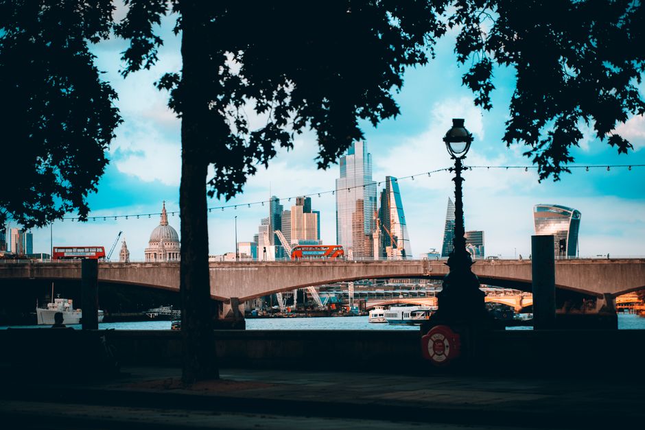 A daytime cityscape view through a park, featuring large, leafy trees in the foreground with dense dark green foliage partially obscuring the scene. Between the trees, there is a vintage-style street lamp with a black metal pole and a glass-encased lantern on top, positioned centrally in the image. In the background, a concrete bridge spans across a river, with boats docked along the water's edge beneath it. Beyond the bridge, the skyline is dominated by modern skyscrapers, including a tall, glass-covered tower with a pointed peak, and other high-rise buildings with reflective glass facades and angular designs. To the left, a historical dome-shaped building with a prominent dome and classical architecture is visible, contrasting with the modern structures. Strings of decorative lights are strung across the park area, hanging above the pathway, which suggests an urban park setting that may be used for leisure or outdoor events, supporting the context of alternative waste handling or removal in city environments thanks to the visible infrastructure. The lighting indicates a partly cloudy sky, with soft, natural light illuminating the scene, highlighting the mix of greenery and diverse architectural styles in the cityscape.