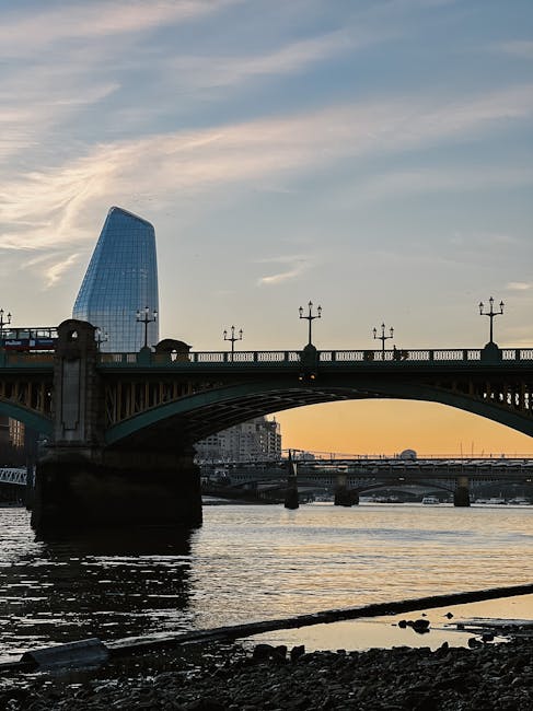 A panoramic view of a bridge spanning a river during sunset, with the City Hall building featuring a distinctive curved glass facade visible in the background. The bridge structure includes ornate lamp posts evenly spaced along the balustrade, and the dark silhouette of the supporting columns contrasts with the warm hues of the setting sun. The river below reflects the sky’s colors, with gentle ripples on the water's surface. The scene captures an urban riverside environment that may relate to alternative waste handling or waste removal services in the area, highlighting the cityscape’s infrastructure against the natural waterway during twilight.